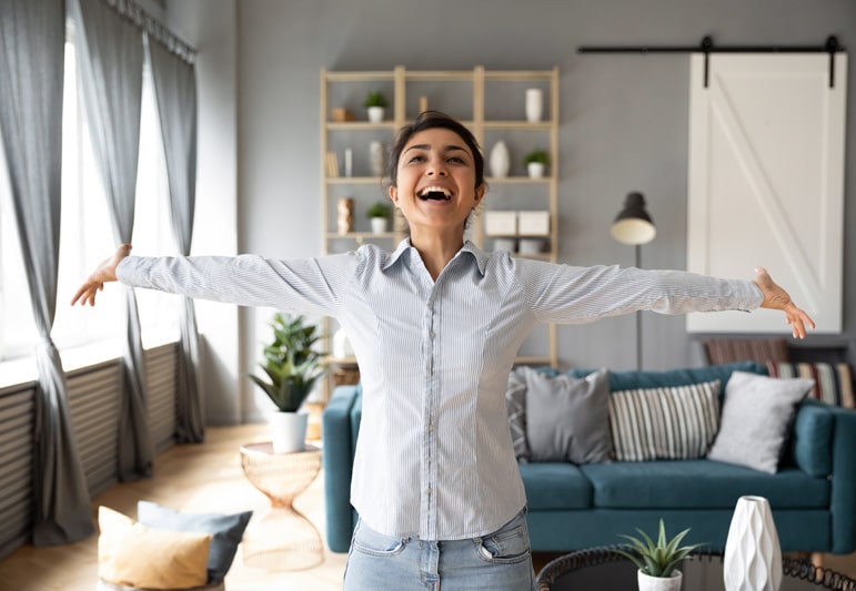 Smiling young woman standing in middle of a living room with her arms outstretched in gratitude and happiness.