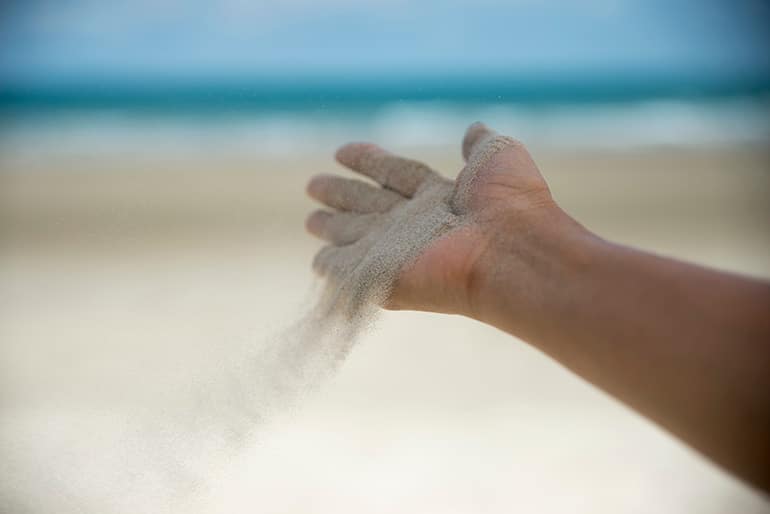 A person at the beach with their outstretched hand letting sand flow into the breeze.