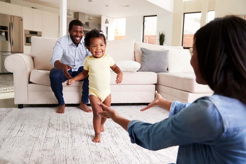 A toddler is taking their first steps from dad to mom in the family living room.