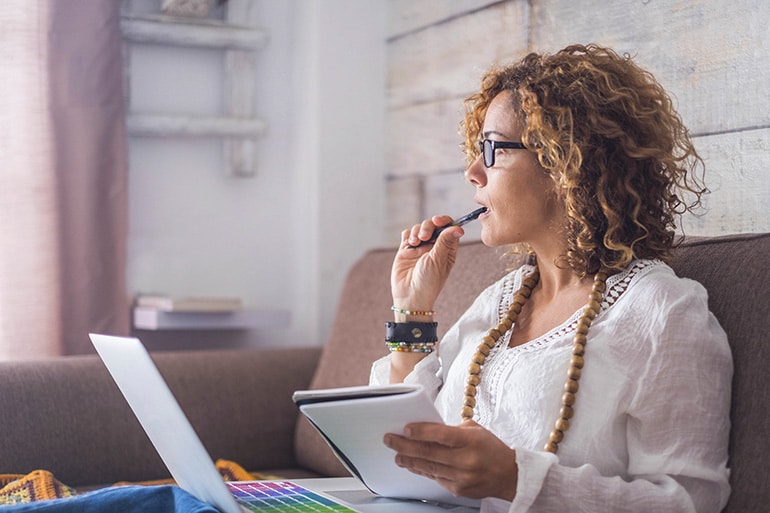 A woman sitting on a couch with a laptop and a notebook holding a pen to her mouth pensively