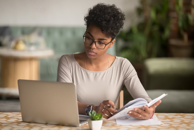 Focused female student looking at laptop while taking notes in a journal.