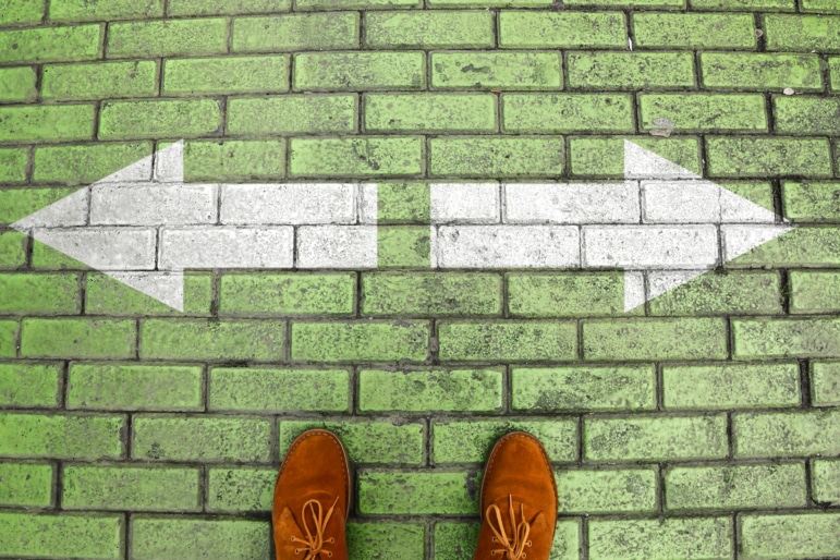 Person's feet in brown suede shoes standing on green brick pavement with white arrows print pointing in two different directions. Two ways to choose making decision which way to go.