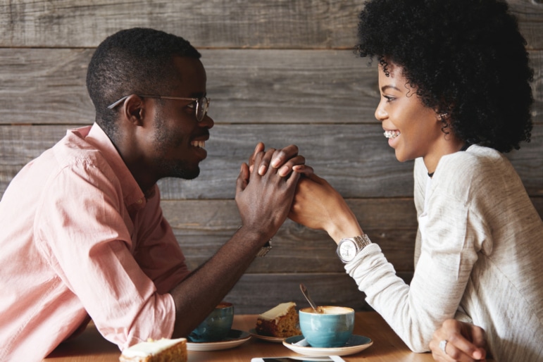 Happy young couple sitting at a coffeeshop holding hands above the table and smiling at each other.