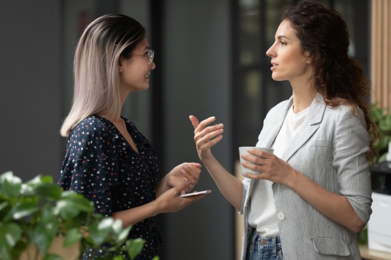 Two smiling young women having a pleasant conversation discussing work during a coffee break in a courtyard.