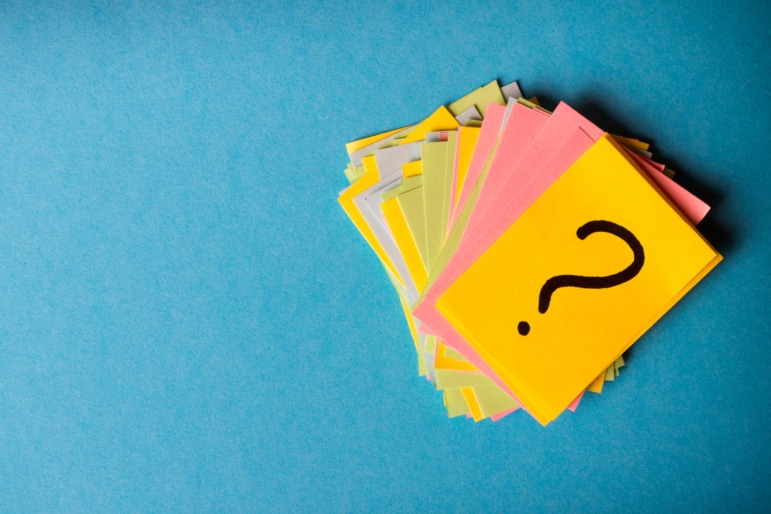 Stacked pile of multicolored cards with a black question mark on top on a light teal blue background.