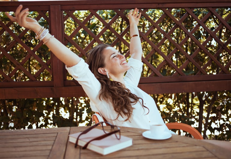 happy modern woman in white shirt with cup of coffee, book and eyeglasses sitting at the table on a covered patio with raised arms rejoicing.