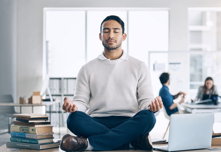 man seated atop his desk in an office space in a meditation pose