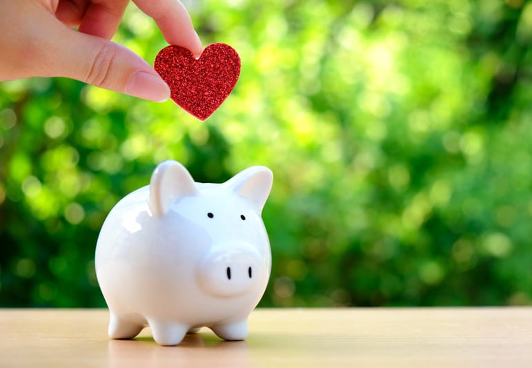 A handholding a small glittery red heart over a white piggy bank with sun drenched green bushes blurred in the background.