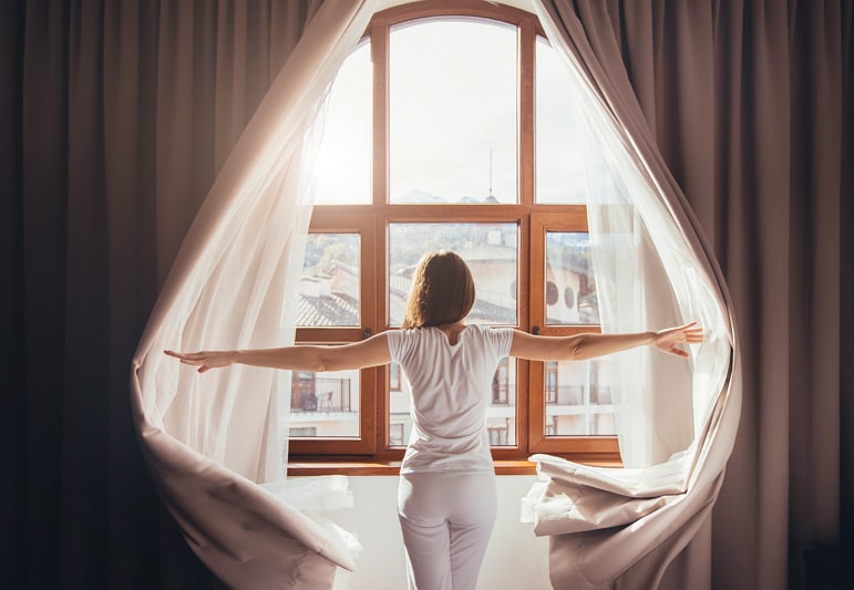 A woman in white pajamas is throwing open heavy curtains to a large window showing a bright sunny city.