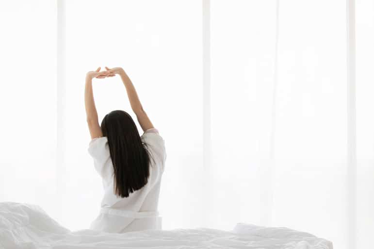 A woman with her back to the camera stretching her arms overhead while sitting on a bed.