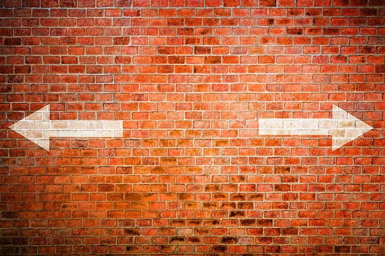Two white arrows facing opposite directions on a brick wall indicating a physician hitting a brick wall and needing to choose burnout or control.