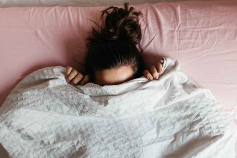 Woman in bed with pink sheets pulling a white blanket over her head.