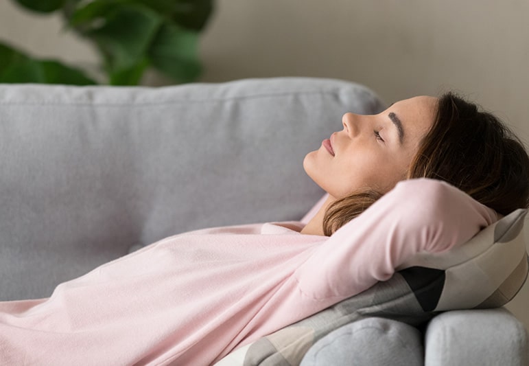 A young woman in a light pink shirt reclines on a light gray couch with her arms behind her head in a relaxed and rested pose with her eyes closed.