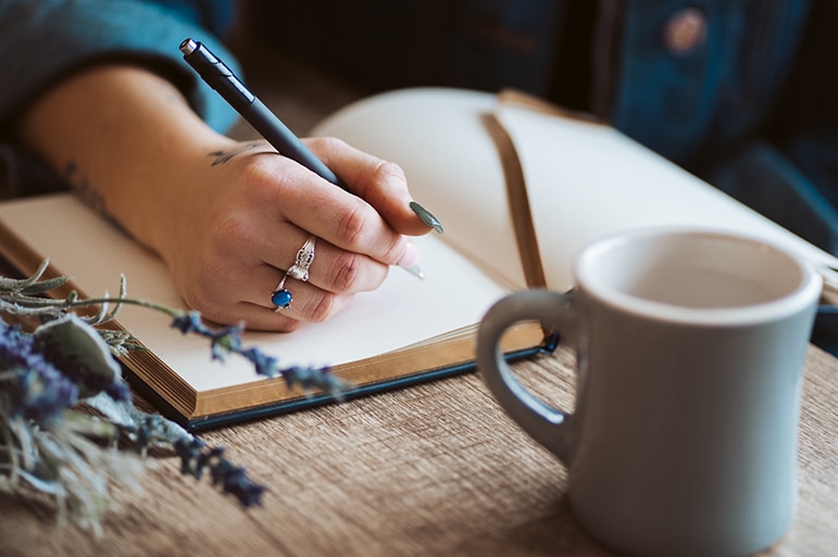 A woman is writing in a journal with a cup of coffee in the foreground.