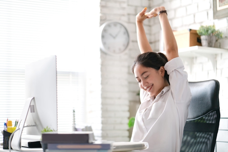 Satisfied clinician stretching her arms over head at her desk in a bright and airy office setting.