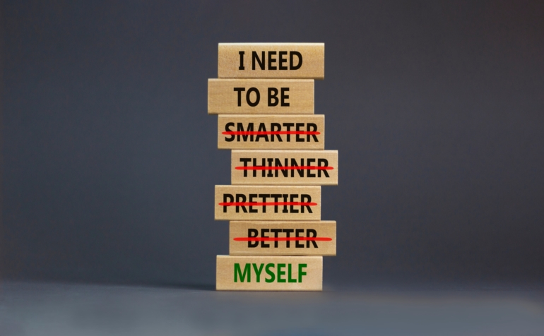 Stacked wooden blocks with I Need To Be on the top, smarter, thinner, better are crossed out and the bottom block says Myself. I need to be myself is the message.