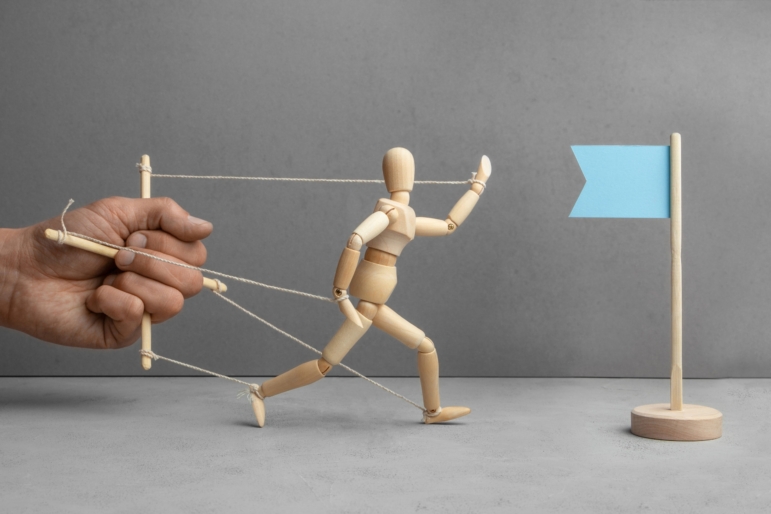 A puppeteer's hand holds the wooden doll back from reaching a flag as symbol of the goal.