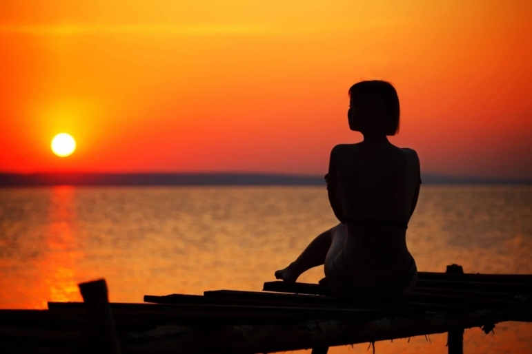 Silhouette of a woman sitting on a wooden pier watching a vibrant orange sunset over a lake.