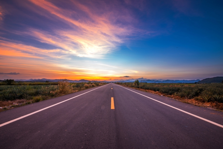 An empty two-lane road stretching straight toward a glowing orange-and-pink sunset horizon, bordered by open fields under a vast sky.