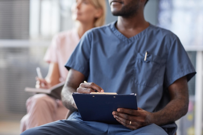 Cropped shot of African American man in blue scrubs keeping records of meeting on clipboard attending lecture in conference room while taking continuing learning course at hospital