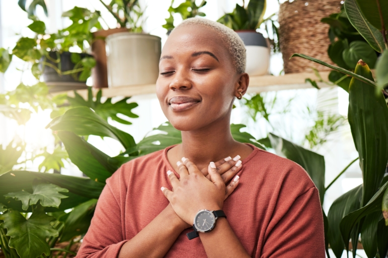 Young Black woman with short-cropped blond hair, eyes closed, hands crossed over chest, wearing a rust-colored top and black watch, surrounded by large green houseplants in bright indoor light practicing mindfulness.