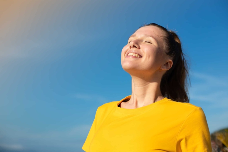 Person standing outdoors with eyes closed, face lifted toward the light, appearing calm and refreshed while pausing to breathe and reset.